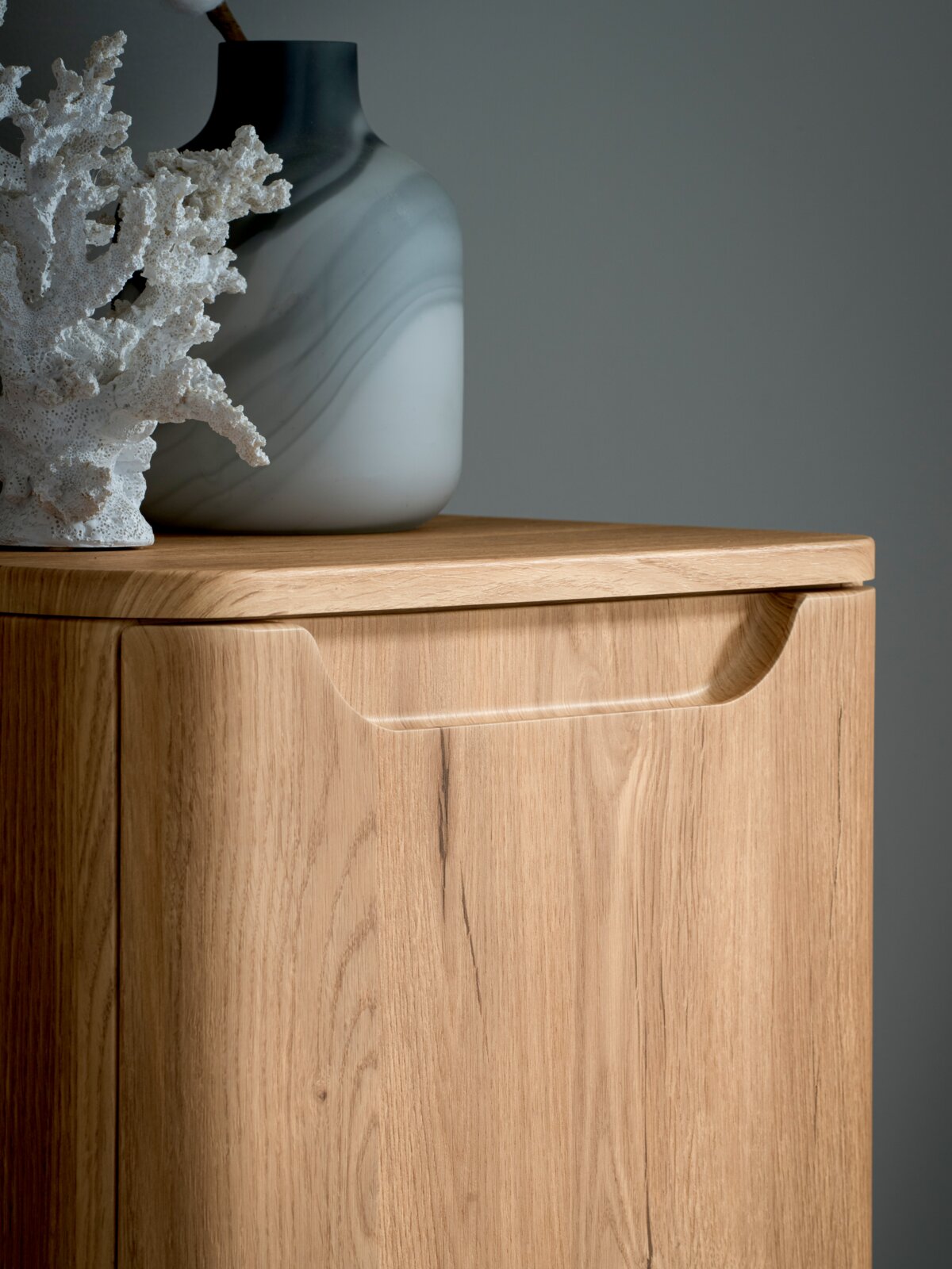 Nahaufnahme eines hellen Holz-Sideboards/Schranks mit integrierter Griffmulde; darauf eine grau-weiß marmorierte Vase und weiße Korallendeko, fotografiert aus leicht seitlicher Frontperspektive.