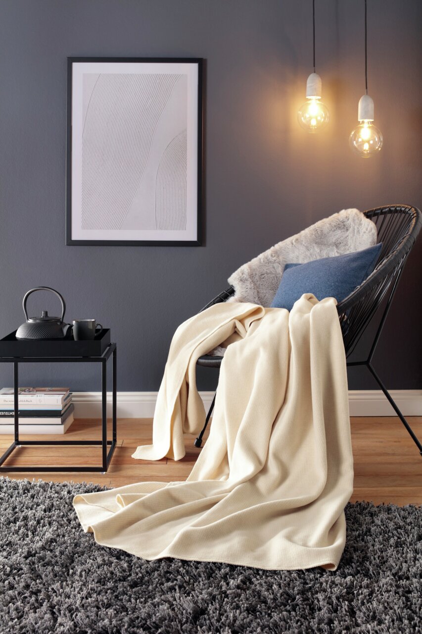 Gemütliche Wohndecke in Cremefarbe auf einem modernen Stuhl in einem stilvoll eingerichteten Wohnzimmer, seitliche Perspektive.