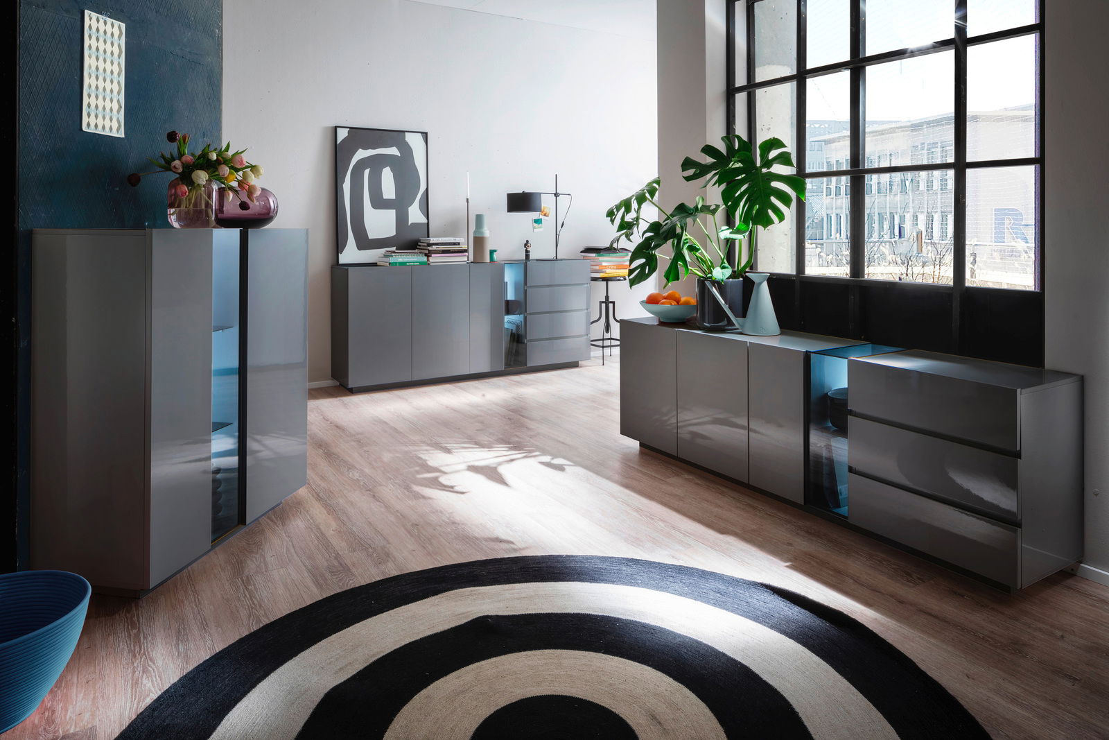 XORA Highboard NAVAN Moderne Wohnzimmermöbel in einem hellen Raum mit großen Fenstern, bestehend aus einem TV-Lowboard, einem XL-Sideboard und einem Highboard in grauem Hochglanz. Perspektive von schräg vorne.