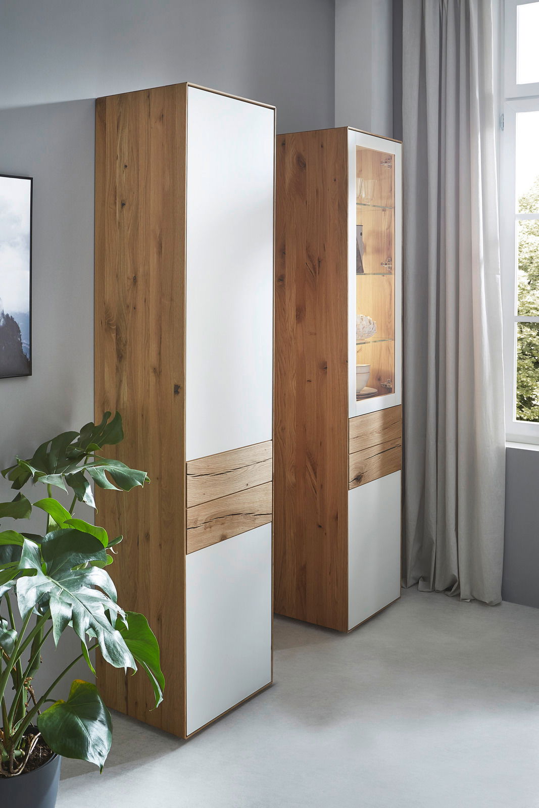 Zwei hohe Stauraumelemente aus Holz mit weißen Fronten, in einem modernen Raum mit einem Fenster und einer Pflanze, seitliche Perspektive.