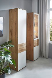 Zwei hohe Stauraumelemente aus Holz mit weißen Fronten, in einem modernen Raum mit einem Fenster und einer Pflanze, seitliche Perspektive.