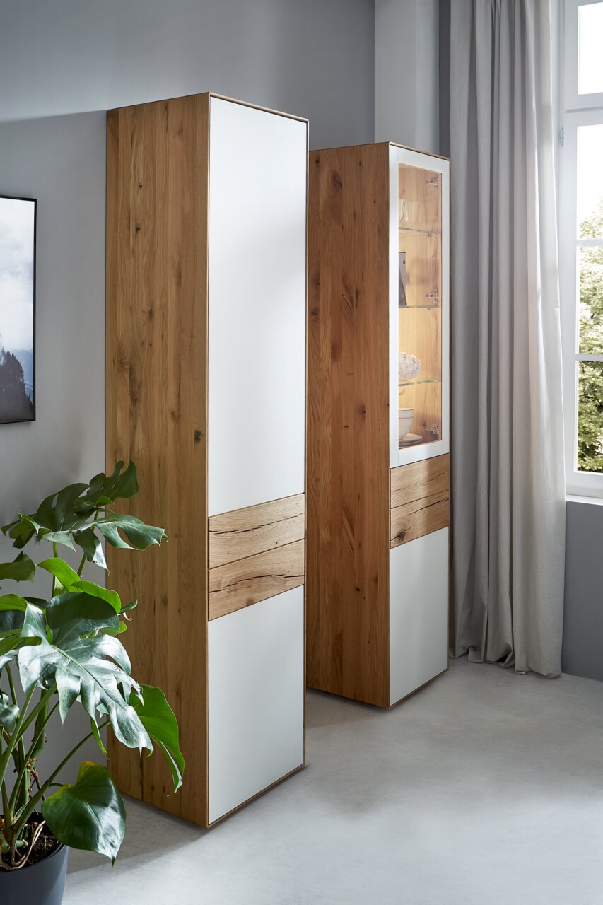 Valnatura Stauraumelement SOLIDA Zwei hohe Stauraumelemente aus Holz mit weißen Fronten, in einem modernen Raum mit einem Fenster und einer Pflanze, seitliche Perspektive.