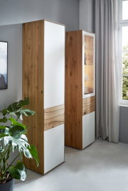 Zwei hohe Stauraumelemente aus Holz mit weißen Fronten, in einem modernen Raum mit einem Fenster und einer Pflanze, seitliche Perspektive.