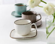 Beige Espressotasse Casa 80ml auf einem passenden Unterteller, im Vordergrund, mit zwei weiteren Tassen in verschiedenen Farben im Hintergrund, seitliche Perspektive.