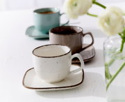 Beige Espressotasse Casa 80ml auf einem passenden Unterteller, im Vordergrund, mit zwei weiteren Tassen in verschiedenen Farben im Hintergrund, seitliche Perspektive.