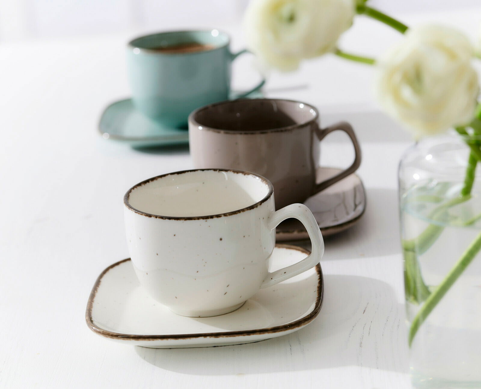 Beige Espressotasse Casa 80ml auf einem passenden Unterteller, im Vordergrund, mit zwei weiteren Tassen in verschiedenen Farben im Hintergrund, seitliche Perspektive.