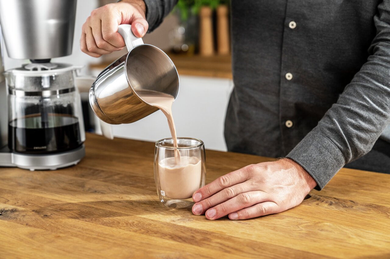 Koffeglas met een inhoud van 200 ml, waaruit iemand koffie inschenkt. Zijaanzicht op een houten werkblad.