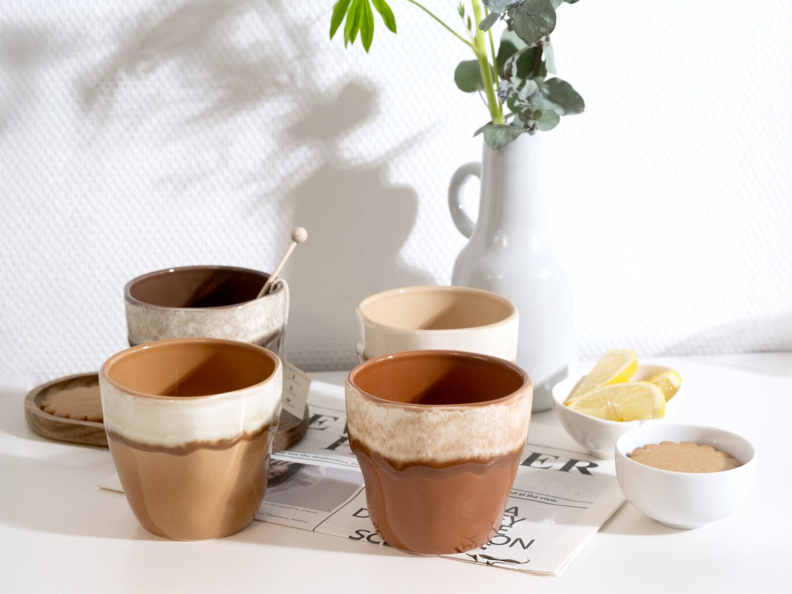 Creatable Teebecher 4-tlg SQUEEZY Drei handgefertigt wirkende Keramikbecher in Braun- und Cremetönen auf weißem Tisch, mit weißer Vase und kleinen Schälchen im Hintergrund, fotografiert aus leicht erhöhter Frontperspektive.