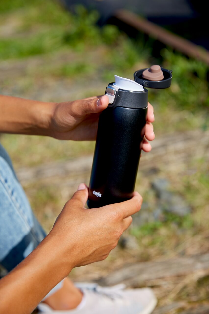 Schwarzer Thermobecher mit geöffnetem Deckel, in der Hand gehalten, im Freien.