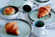 Stilvolles Keramikgeschirr-Set in Weiß mit blaugrüner, gesprenkelter Glasur (Teller, Schalen und Tassen) mit Croissants und Zimtschnecke arrangiert; Perspektive: leicht von oben in schräger Draufsicht.