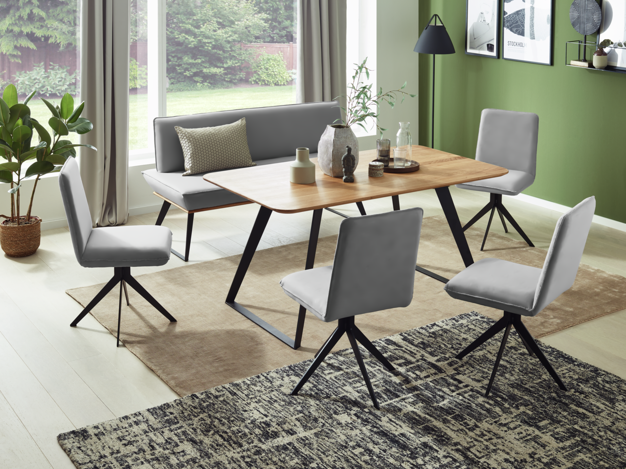 Voleo Esstisch MALIBU Moderner Essbereich mit einem Esstisch aus Holz und schwarzen Metallbeinen, umgeben von grauen Stühlen und einer Eckbank, aus der Perspektive des Raumes mit Blick auf große Fenster.