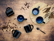 Draufsicht von oben auf schwarze Kaffeetassen mit blau glasiertem Innenraum auf rustikalem Holztisch, mit Kaffeebohnen, Holzbrett und Löffel