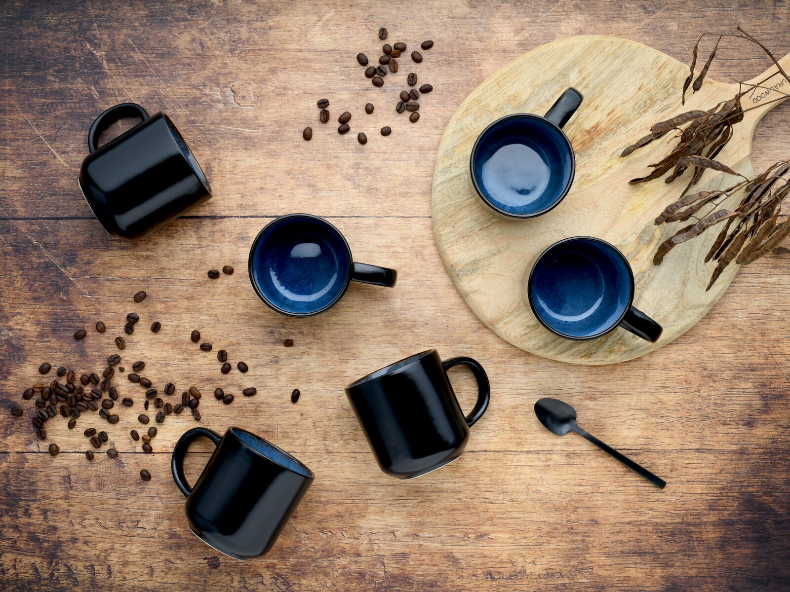 Draufsicht von oben auf schwarze Kaffeetassen mit blau glasiertem Innenraum auf rustikalem Holztisch, mit Kaffeebohnen, Holzbrett und Löffel