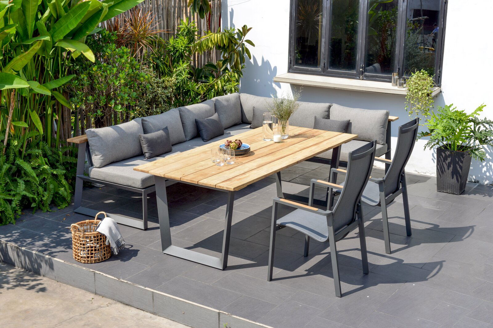 Moderner Gartentisch mit Holzplatte und Metallgestell, umgeben von grauen Stühlen und einer Eck-Sitzbank auf einer Terrasse, seitliche Perspektive.