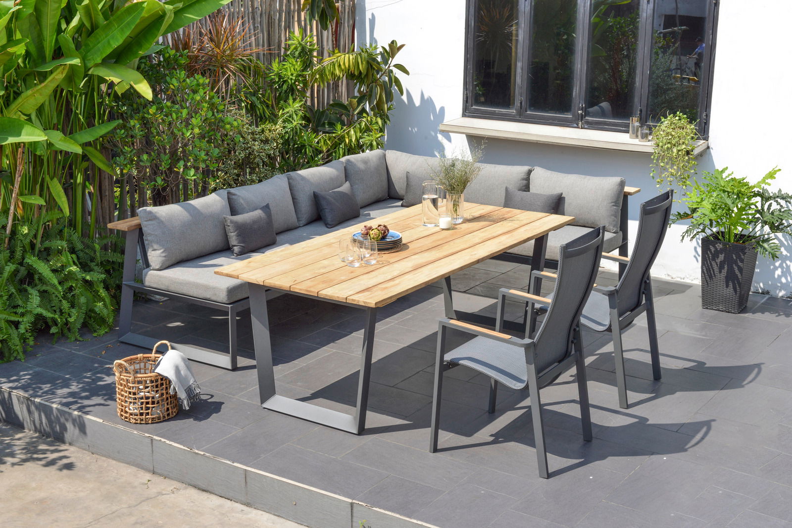 Moderner Gartentisch mit Holzplatte und Metallgestell, umgeben von grauen Stühlen und einer Eck-Sitzbank auf einer Terrasse, seitliche Perspektive.