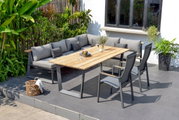 Moderner Gartentisch mit Holzplatte und Metallgestell, umgeben von grauen Stühlen und einer Eck-Sitzbank auf einer Terrasse, seitliche Perspektive.