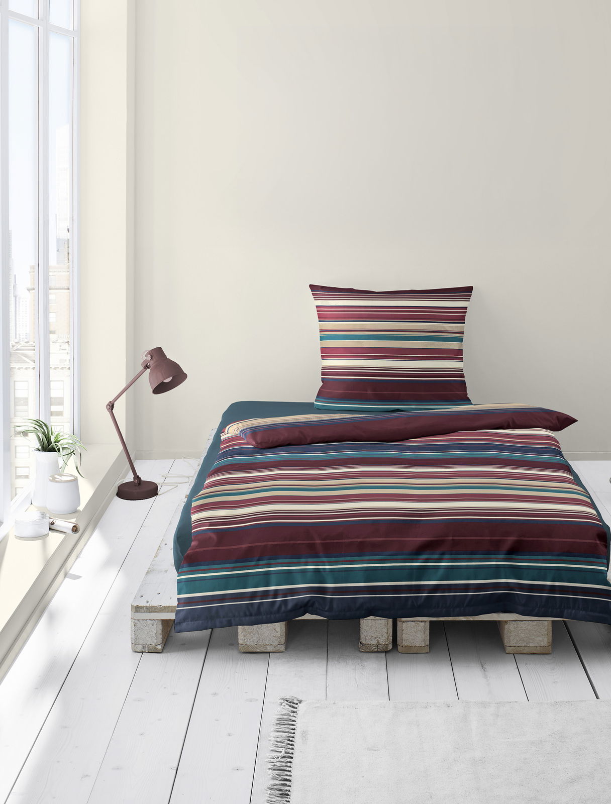 Bettwäsche-Set mit bunten Streifen auf einem Palettenbett, fotografiert aus der Vorderansicht in einem hellen Raum mit Fenster und Stehlampe.