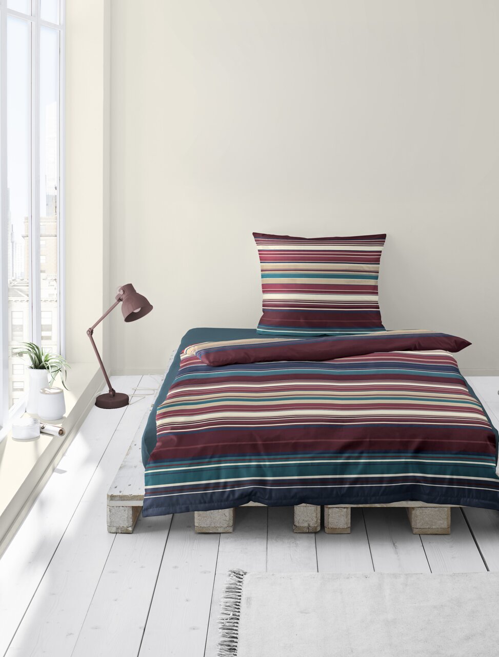 Bettwäsche-Set mit bunten Streifen auf einem Palettenbett, fotografiert aus der Vorderansicht in einem hellen Raum mit Fenster und Stehlampe.