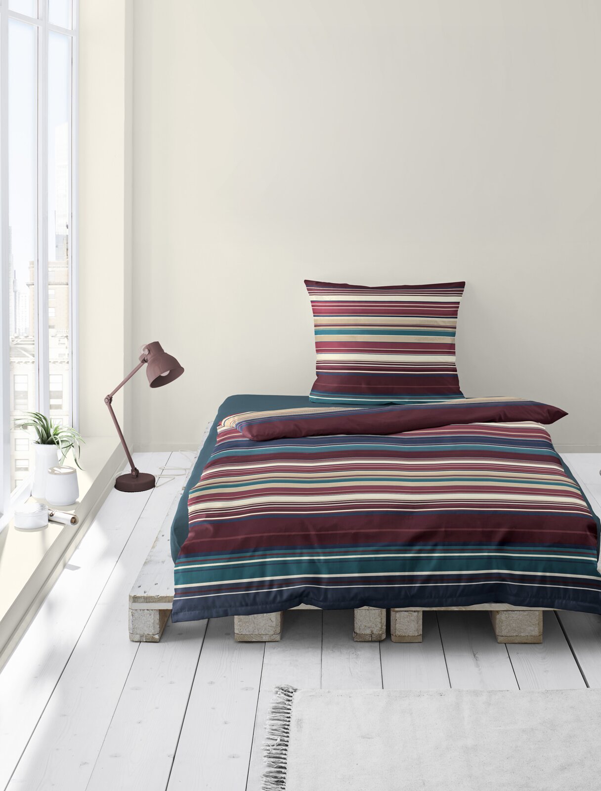 Beddengoedset met kleurrijke strepen op een palletbed, gefotografeerd van voren in een lichte kamer met een raam en een staande lamp.