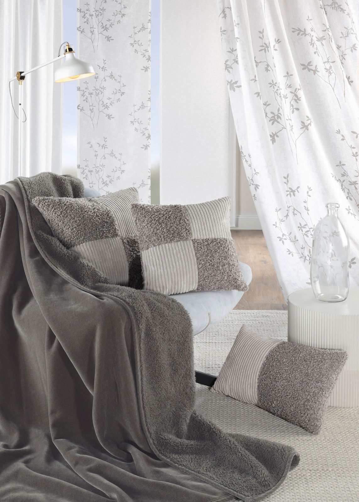 GÖZZE Fertigvorhang VICTORIA Wohnzimmer mit grauen Kissen und Decke auf einem Sofa, im Hintergrund weiße Vorhänge mit floralem Muster, seitliche Perspektive.