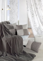 Wohnzimmer mit grauen Kissen und Decke auf einem Sofa, im Hintergrund weiße Vorhänge mit floralem Muster, seitliche Perspektive.