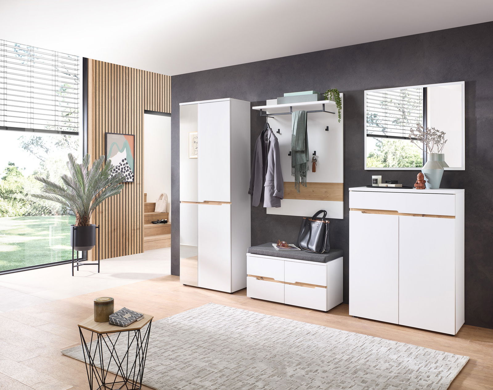 XORA Garderobenset 5-tlg. MEMPHIS Moderne Garderobenset in einem hellen Raum mit großem Fenster, bestehend aus einem hohen Schrank, einem Garderobenpaneel mit Haken und Ablage, einer Sitzbank mit Stauraum und einem Spiegel. Perspektive von vorne.