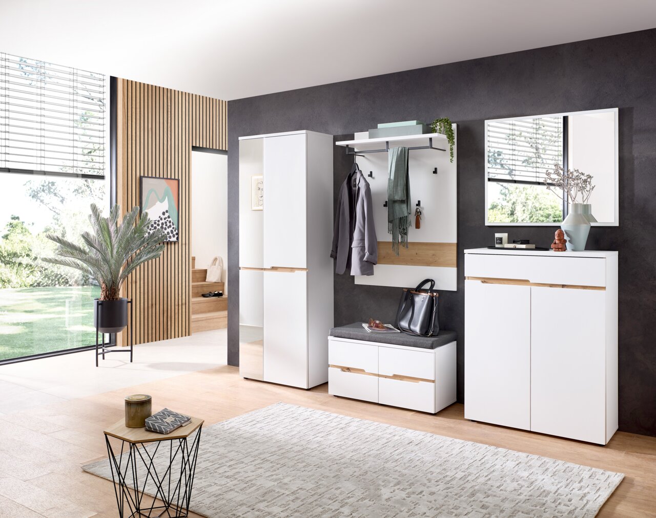 Moderne Garderobenset in einem hellen Raum mit großem Fenster, bestehend aus einem hohen Schrank, einem Garderobenpaneel mit Haken und Ablage, einer Sitzbank mit Stauraum und einem Spiegel. Perspektive von vorne.