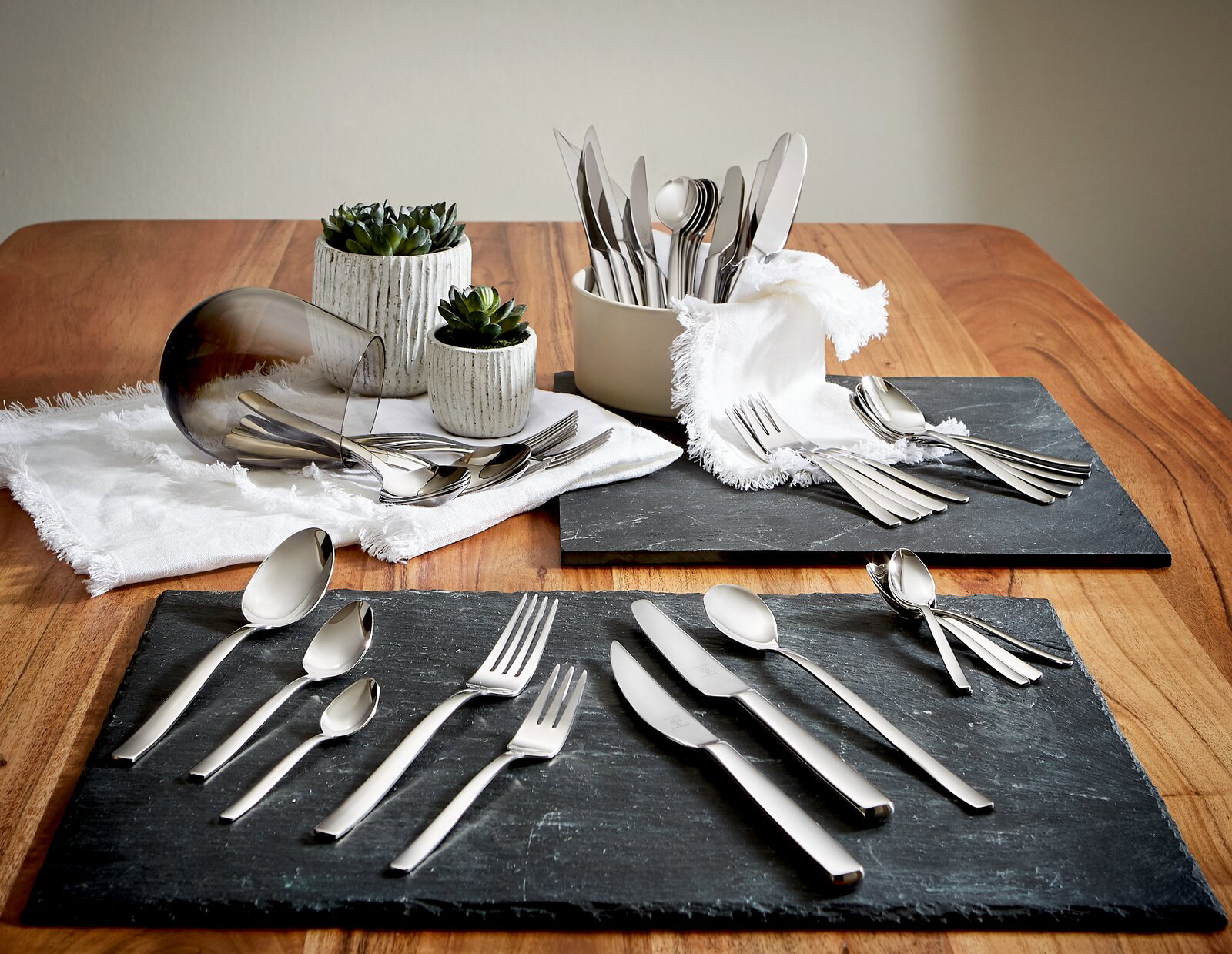 Elegantes 48-teiliges Besteckset auf einem Holztisch, präsentiert auf Schieferplatten mit dekorativen Pflanzen im Hintergrund. Perspektive von oben.