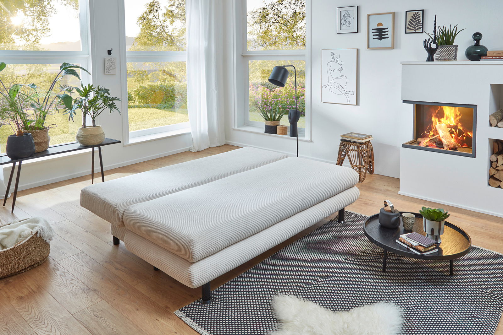 Modernes Funktionssofa Bern in einem hellen Wohnzimmer mit großen Fenstern, Pflanzen und einem Kamin, aus der Seitenperspektive fotografiert.