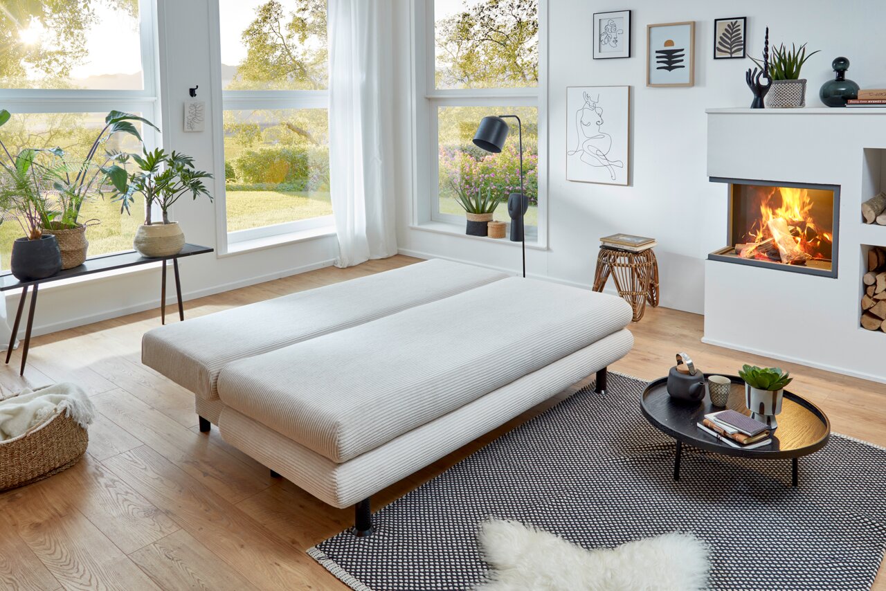 Modernes Funktionssofa Bern in einem hellen Wohnzimmer mit großen Fenstern, Pflanzen und einem Kamin, aus der Seitenperspektive fotografiert.