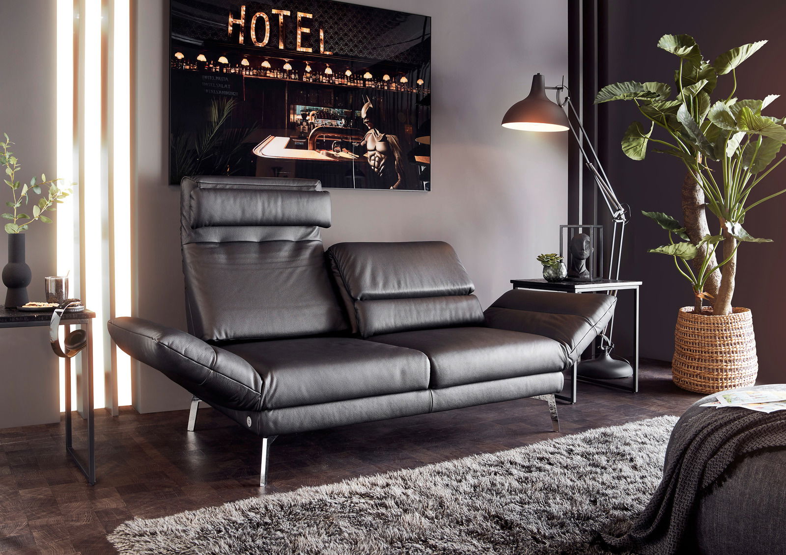 Modernes, schwarzes Ledersofa mit verstellbaren Kopfstützen, seitlicher Perspektive, in stilvollem Wohnzimmer mit Stehlampe und Pflanzen.
