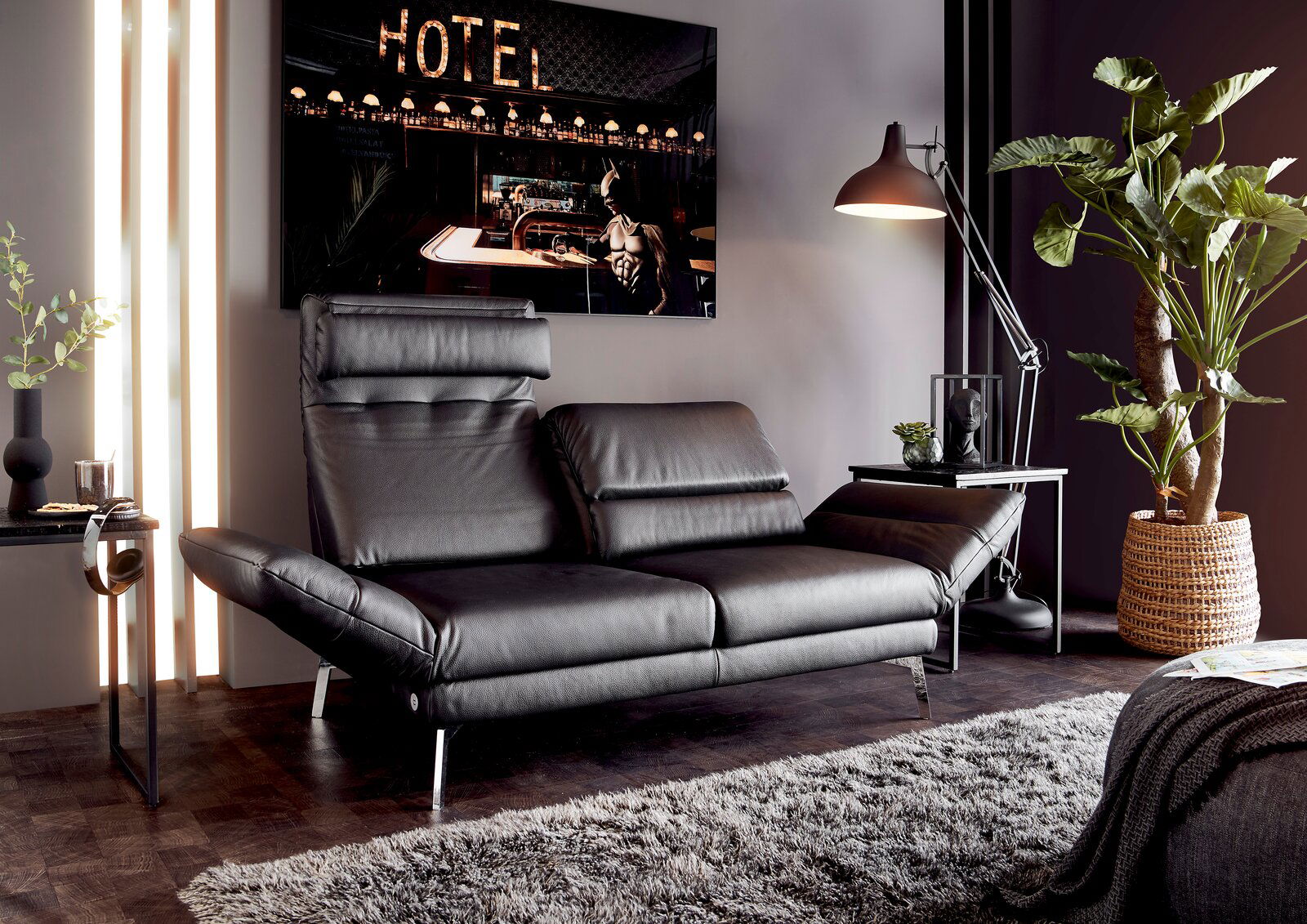 Modernes, schwarzes Ledersofa mit verstellbaren Kopfstützen, seitlicher Perspektive, in stilvollem Wohnzimmer mit Stehlampe und Pflanzen.