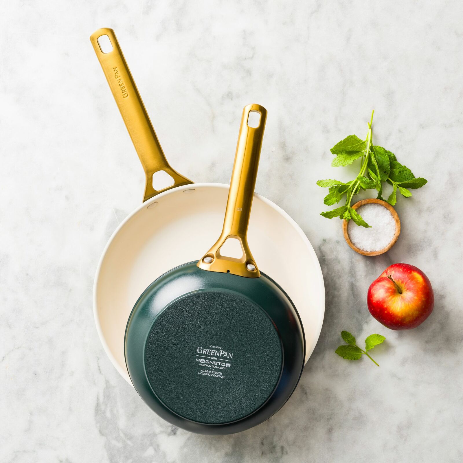 Zwei Bratpfannen-Set mit goldenen Griffen, eine Pfanne liegt in der anderen, von oben fotografiert, neben frischen Kräutern, Salz und einem Apfel auf einer Marmoroberfläche.