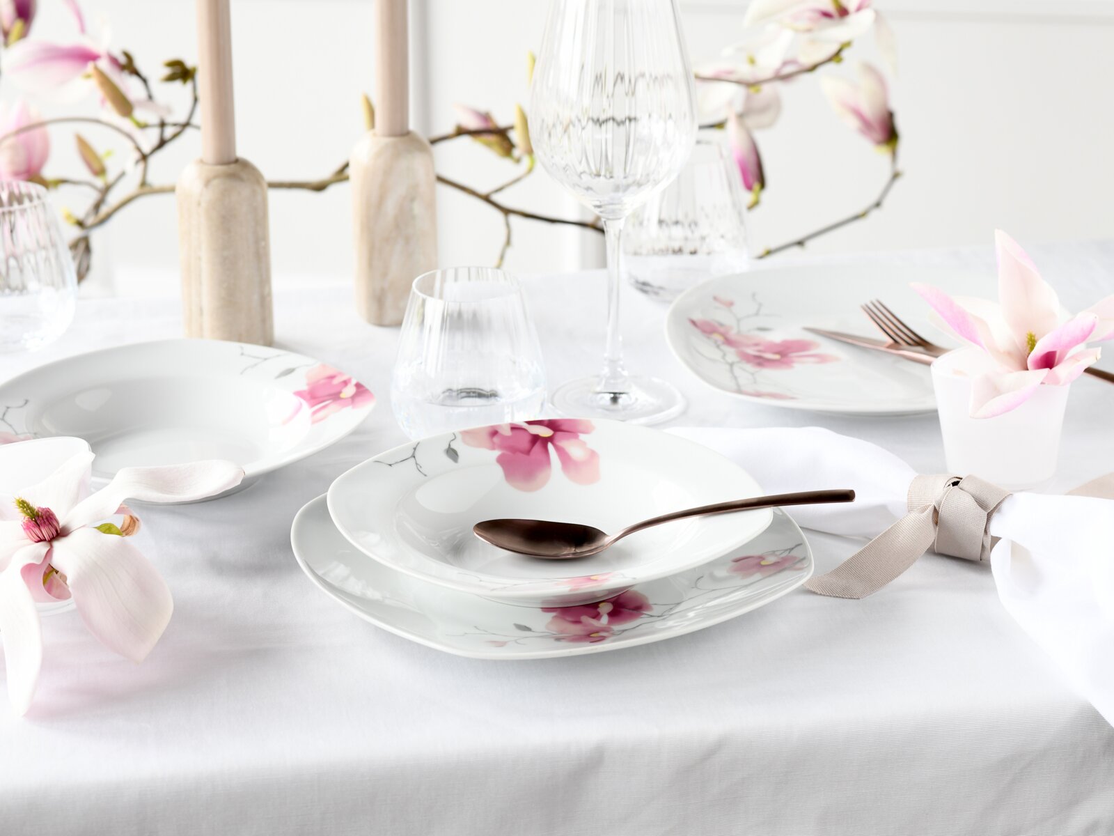 Frühlingshaft gedeckter Tisch mit weißem Porzellan-Geschirrset mit rosa Magnolienblüten-Motiv, Glaswaren und Besteck auf weißer Tischdecke, Perspektive: leicht von oben in schräger Frontansicht