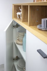 Nahaufnahme einer Kommode mit geöffneter Tür, die Tassen und Teller zeigt. Oben sind offene Fächer mit einem dekorativen Hirsch. Perspektive von der Seite.