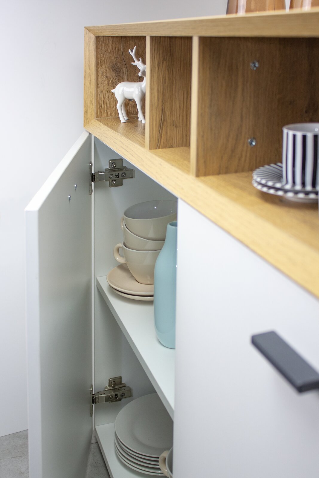 Nahaufnahme einer Kommode mit geöffneter Tür, die Tassen und Teller zeigt. Oben sind offene Fächer mit einem dekorativen Hirsch. Perspektive von der Seite.
