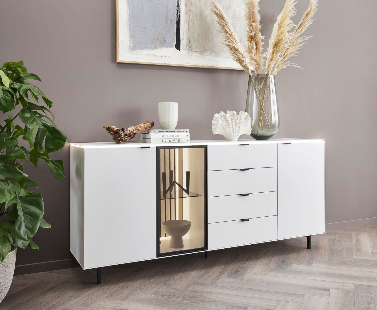 Dieter Knoll Dressoir RIANI Modernes weißes Sideboard mit Schubladen und Glastür, dekoriert mit Büchern, Vase und Pflanzen, seitliche Perspektive.