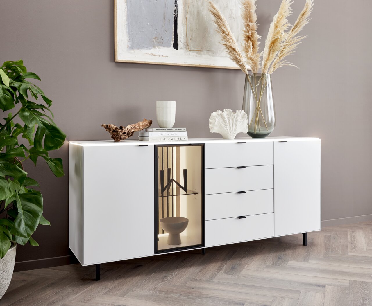 Modernes weißes Sideboard mit Schubladen und Glastür, dekoriert mit Büchern, Vase und Pflanzen, seitliche Perspektive.