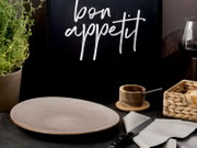 Beiger, gesprenkelter Keramikteller auf dunklem Tisch vor schwarzer Tafel mit Schriftzug „bon appetit“, seitliche Frontansicht mit leichtem Blick von oben; daneben Holzschälchen, Messer, Serviette, Kräutertopf und Brotkorb.