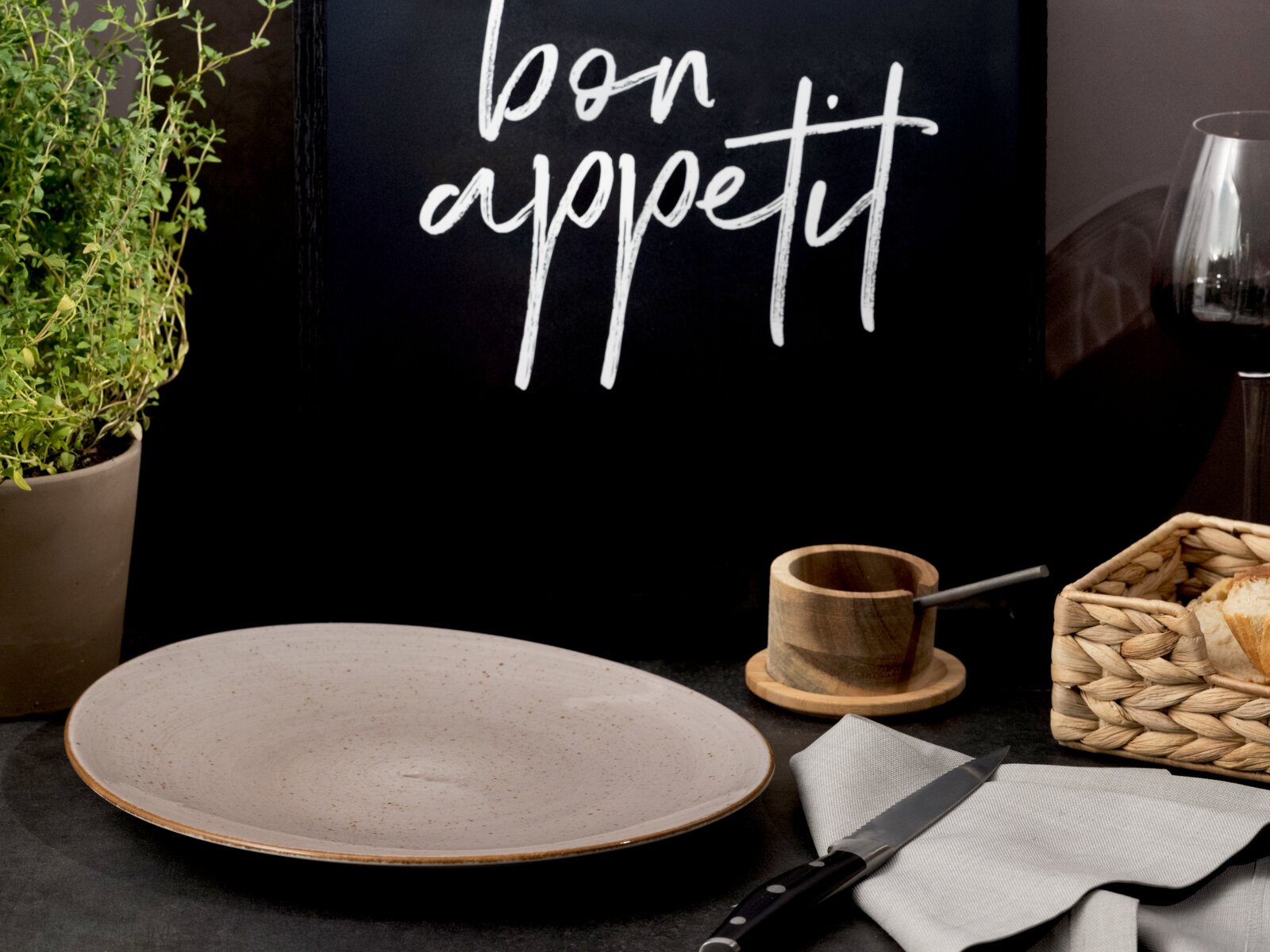 Beiger, gesprenkelter Keramikteller auf dunklem Tisch vor schwarzer Tafel mit Schriftzug „bon appetit“, seitliche Frontansicht mit leichtem Blick von oben; daneben Holzschälchen, Messer, Serviette, Kräutertopf und Brotkorb.