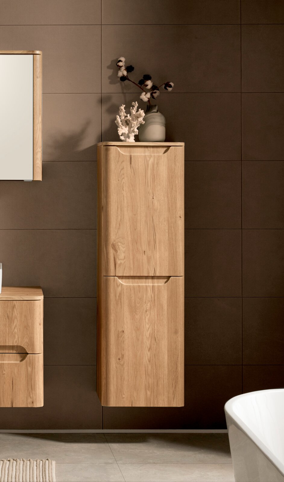 Hochformatiger, wandhängender Bad-Hochschrank aus hellem Holz mit zwei grifflosen Fronten vor dunkelgrau gefliester Wand, frontal aus leichter Seitenperspektive fotografiert; oben dekoriert mit Vase und Baumwollzweigen.