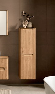 Hochformatiger, wandhängender Bad-Hochschrank aus hellem Holz mit zwei grifflosen Fronten vor dunkelgrau gefliester Wand, frontal aus leichter Seitenperspektive fotografiert; oben dekoriert mit Vase und Baumwollzweigen.