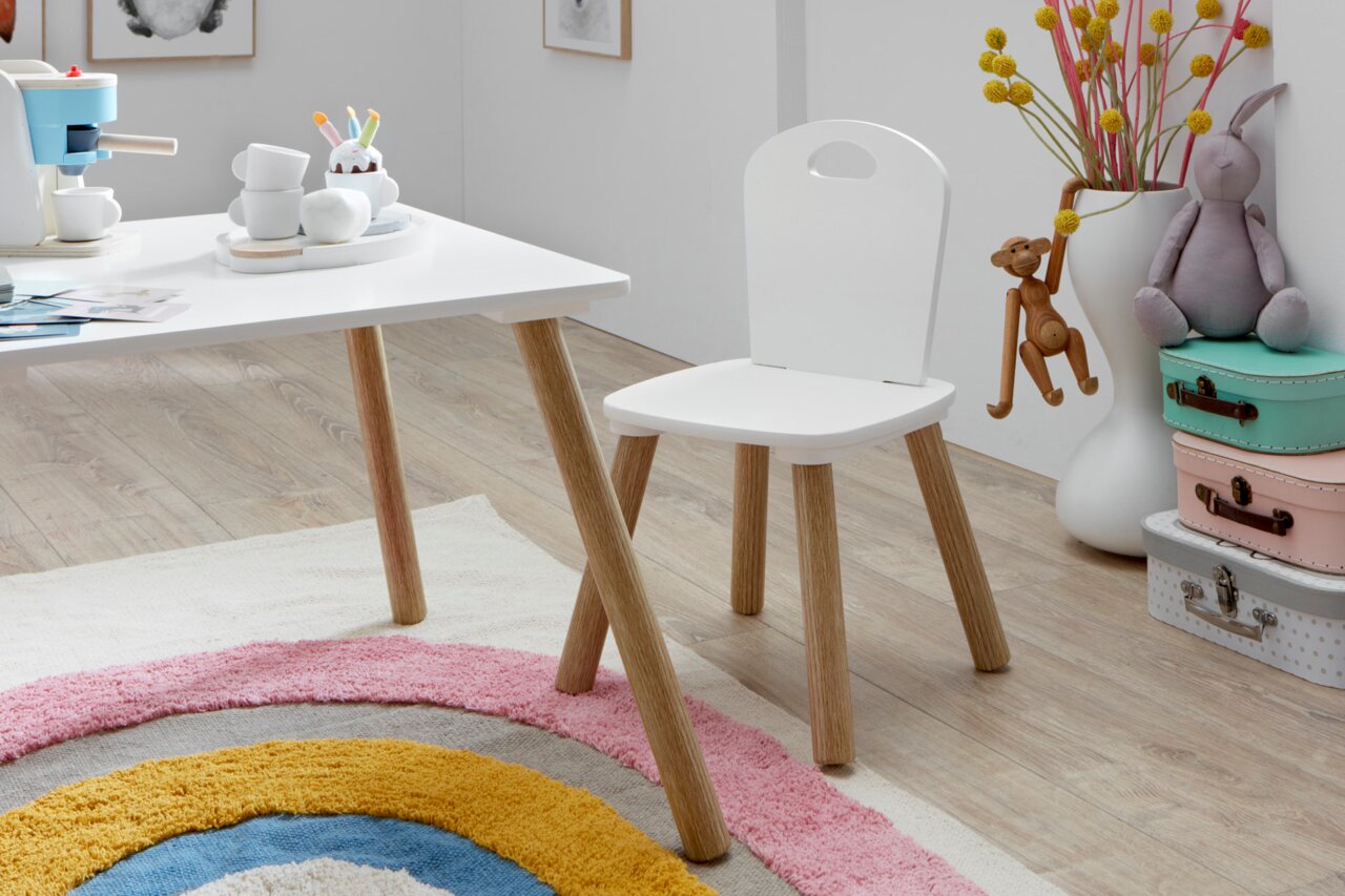 Weißer Kinderstuhl mit Holzbeinen neben passendem weißem Tisch im Kinderzimmer, seitliche Perspektive auf Sitzfläche und Rückenlehne