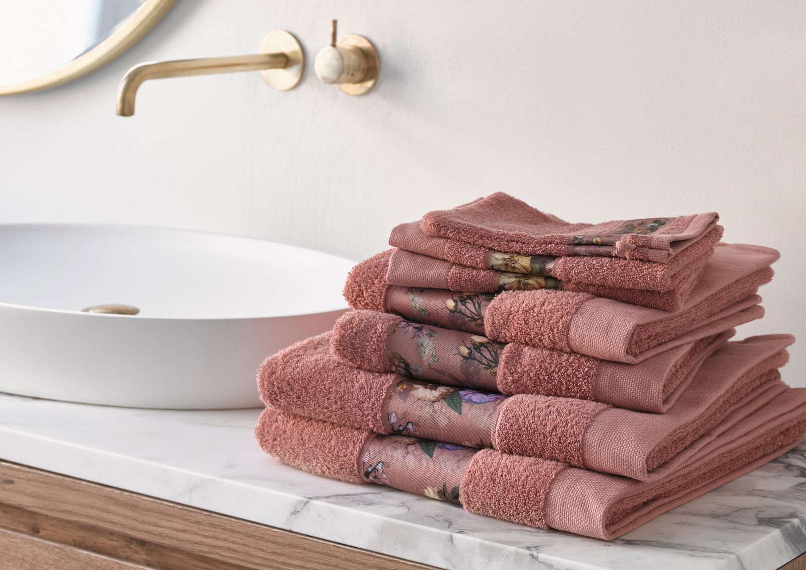 Ein Stapel rosafarbener Handtücher mit floralen Mustern, seitlich auf einem Marmortisch neben einem modernen Waschbecken arrangiert.