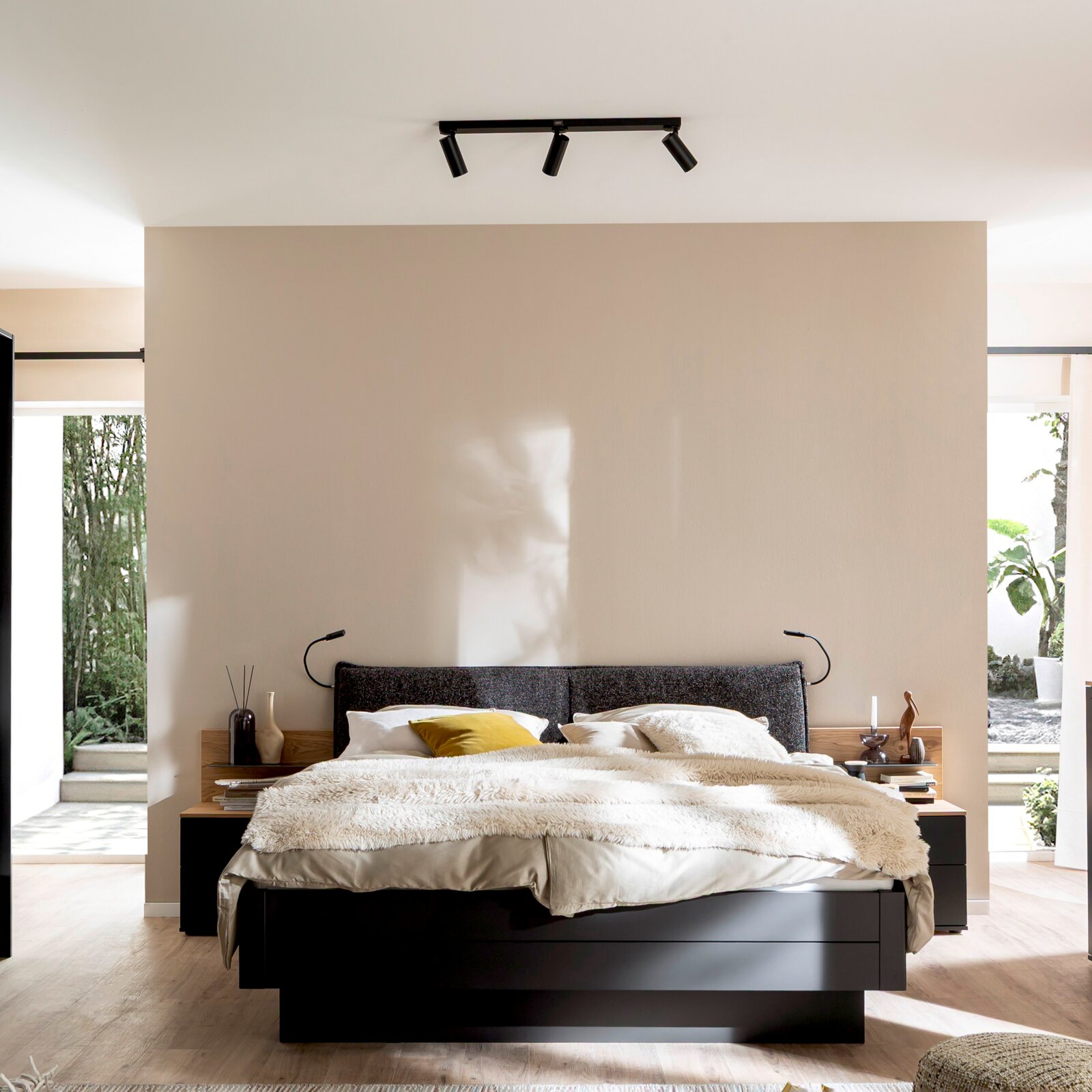 Modernes Schlafzimmer mit Doppelbett und dunklem Bettrahmen, Stoff-Kopfteil, heller Bettwäsche und Decken-Spotleiste an der Decke; frontale Ansicht auf das Bett