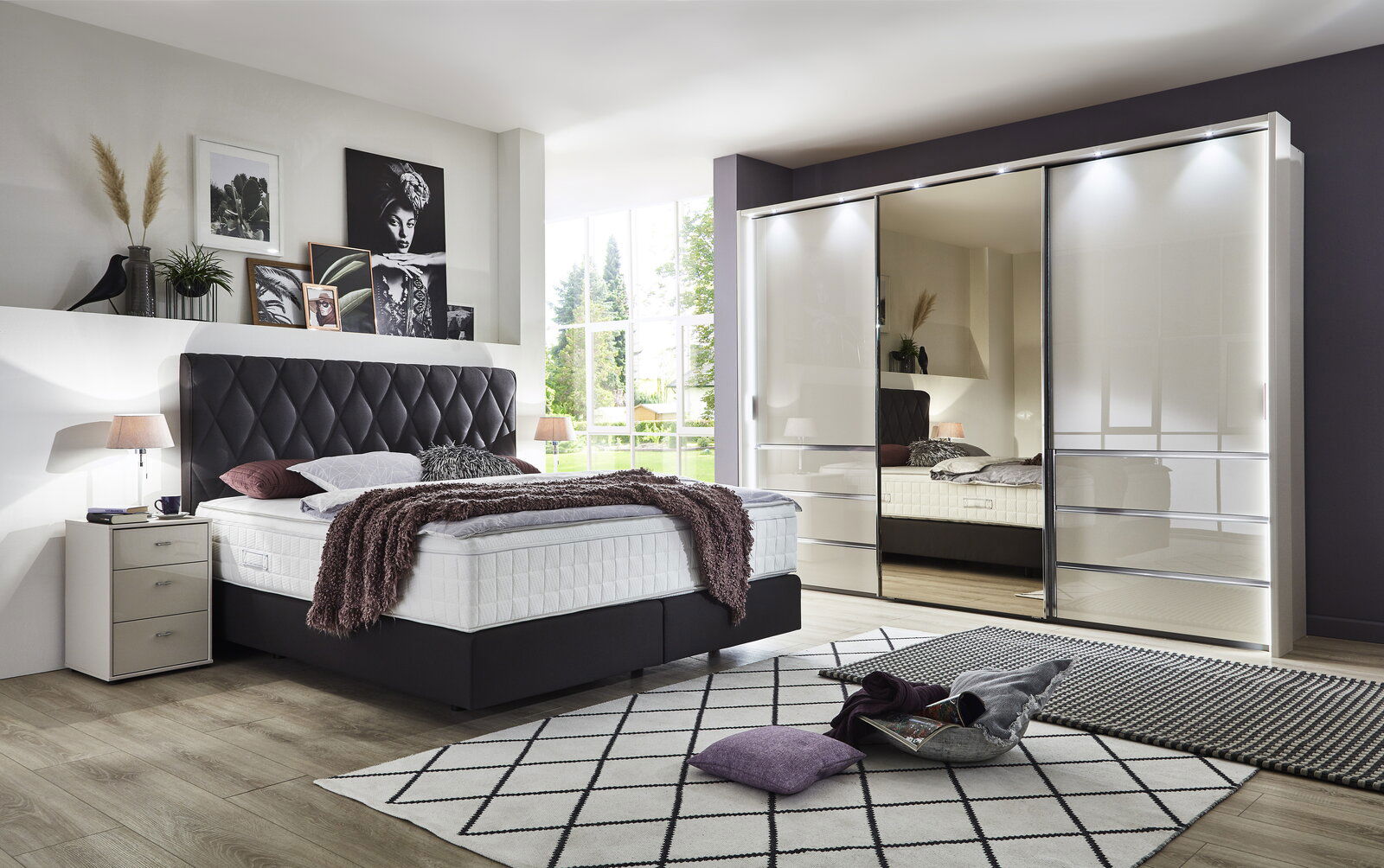 Schlafzimmer mit modernem Schwebetürenschrank MALIBU in Hochglanzoptik, links ein Bett mit gepolstertem Kopfteil, Perspektive von vorne.