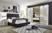 Schlafzimmer mit modernem Schwebetürenschrank MALIBU in Hochglanzoptik, links ein Bett mit gepolstertem Kopfteil, Perspektive von vorne.