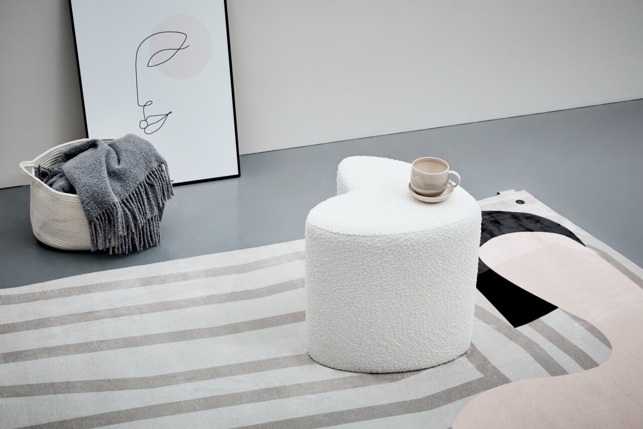 Moderner weißer Hocker in Herzform, seitliche Perspektive, auf einem gemusterten Teppich mit einer Tasse darauf.