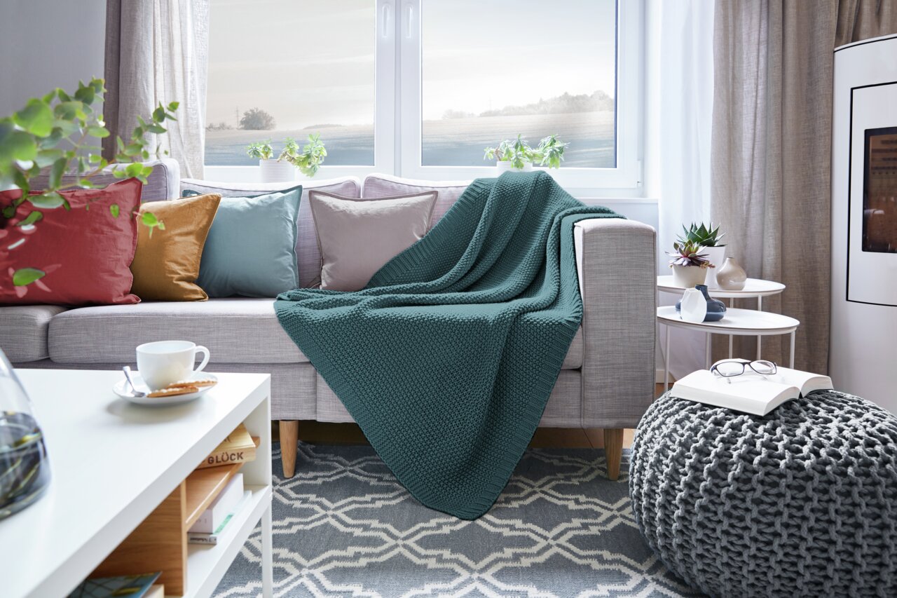 Gemütliches Wohnzimmer mit einer grünen Strickdecke auf einem grauen Sofa, umgeben von bunten Kissen und einem modernen Beistelltisch. Perspektive von vorne.