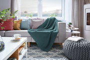 Gemütliches Wohnzimmer mit einer grünen Strickdecke auf einem grauen Sofa, umgeben von bunten Kissen und einem modernen Beistelltisch. Perspektive von vorne.